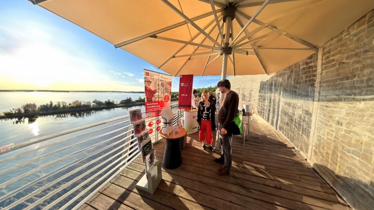 Zwei Menschen unterhalten sich an einem Ausstellungsstand. Über ihnen ist ein Schirm aufgespannt. Links im Bild ist ein See und rechts eine Ziegelsteinwand.