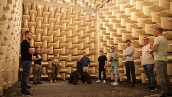 Eine Gruppe von Menschen steht in einem Raum, welcher mit Lautsprechern ausgedeckt ist.