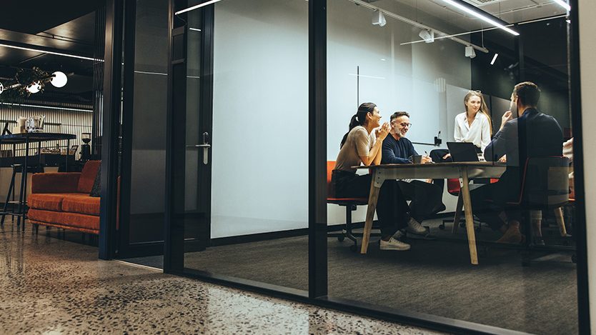 Menschen arbeiten an einem Tisch zusammen. Dabei sitzen sie in einem abgetrennten Bereich aus Glas.