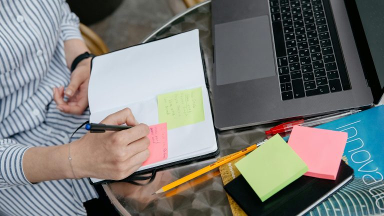 Eine Person sitzt an einem Schreibtisch. Darauf ein Laptop und Notizzettel. Die Person macht handschriftliche Noitzen