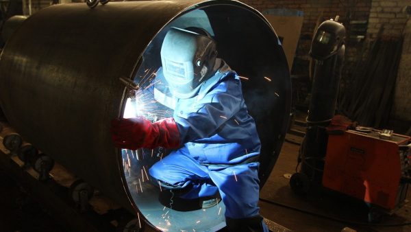 KI in Produktionsunternehmen Person im Schutzanzug sitzt mit Schweißermaske in einer Röhre und schweißt.