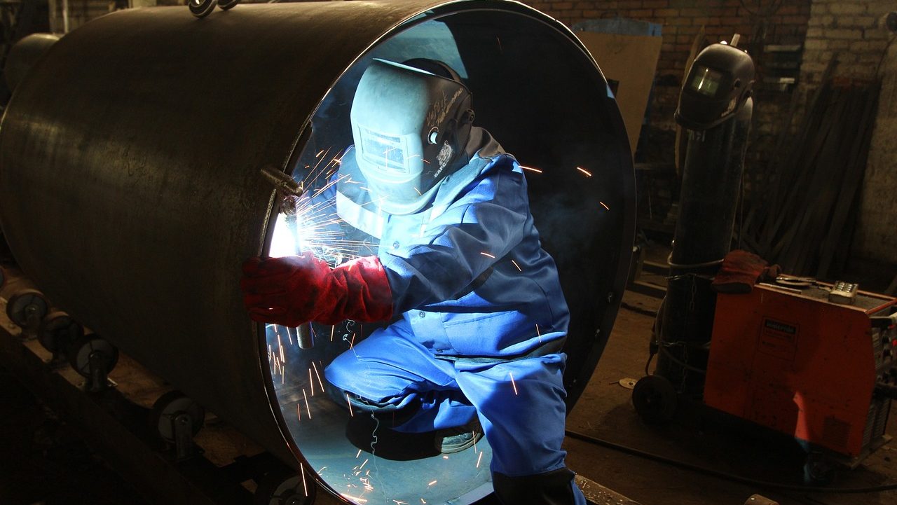 KI in Produktionsunternehmen Person im Schutzanzug sitzt mit Schweißermaske in einer Röhre und schweißt.