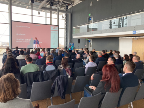 Publikum sitzt vo einem kleinen Podium wo jemand etwas zu KI-Mittelstandstag vorträgt