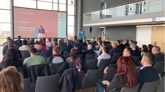 Publikum sitzt vo einem kleinen Podium wo jemand etwas zu KI-Mittelstandstag vorträgt