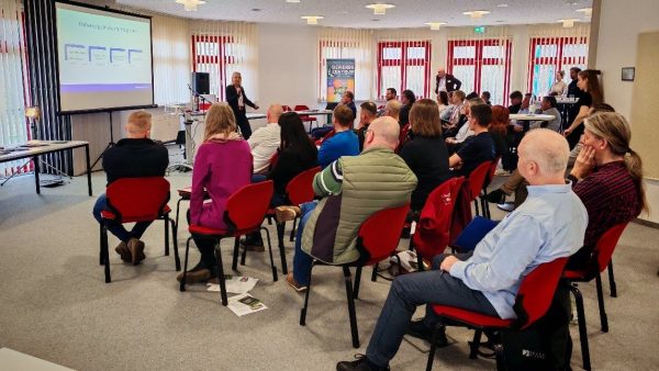 Publikum Viele Menschen sitzen auf Stühlen und schauen auf eine Beamerleinwand, die eine Präsentation zeigt