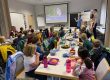 Eine Gruppe von Grundschulkindern sitzt an langen Tischen in einem Klassenraum und schaut nach vorne zu drei Erwachsenen, die vor einer Leinwand präsentieren. Auf den Tischen stehen Brotdosen, Trinkflaschen und Obstteller. Die Kinder wirken aufmerksam und essen gleichzeitig ihre Pausenmahlzeit