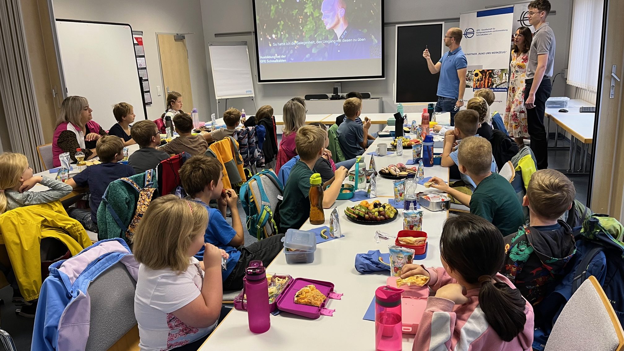 Innovation zum anfassen Eine Gruppe von Grundschulkindern sitzt an langen Tischen in einem Klassenraum und schaut nach vorne zu drei Erwachsenen, die vor einer Leinwand präsentieren. Auf den Tischen stehen Brotdosen, Trinkflaschen und Obstteller. Die Kinder wirken aufmerksam und essen gleichzeitig ihre Pausenmahlzeit
